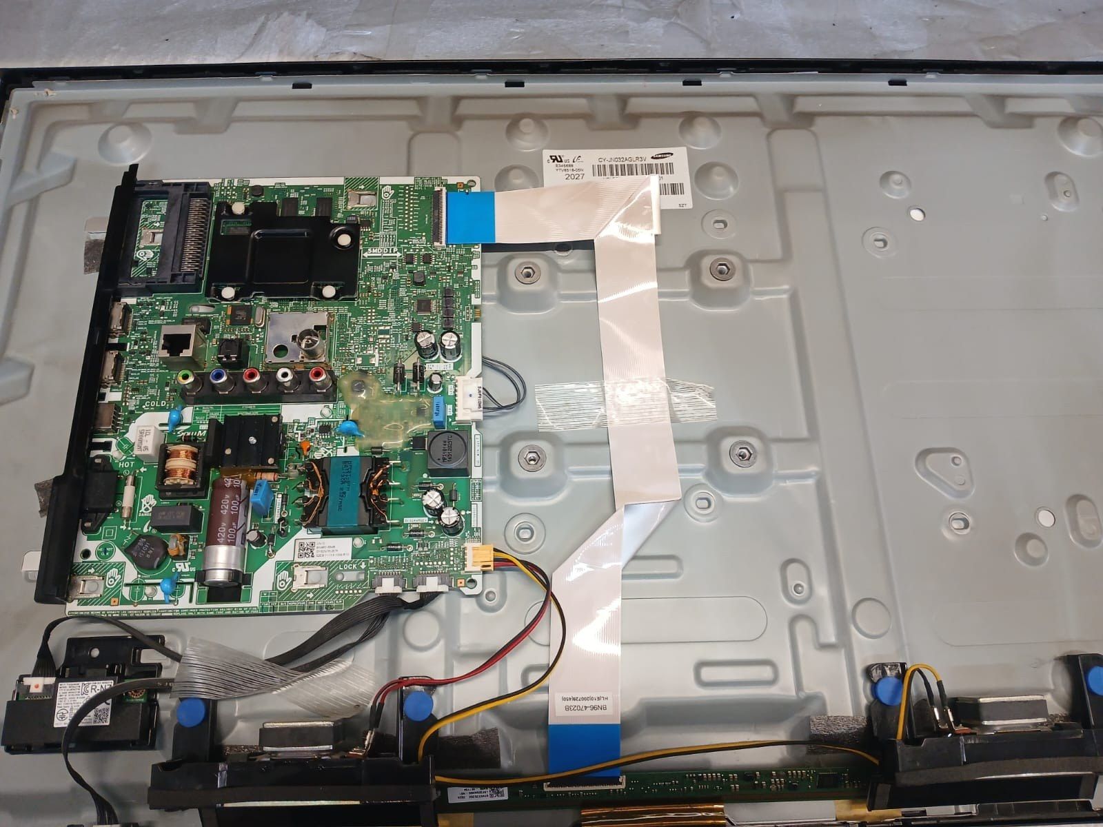 Televizor Samsung desfăcut pe banc: panoul scos din carcasă, placa de bază și sursa de alimentare la vedere, cu cablurile panglică și difuzoarele alături