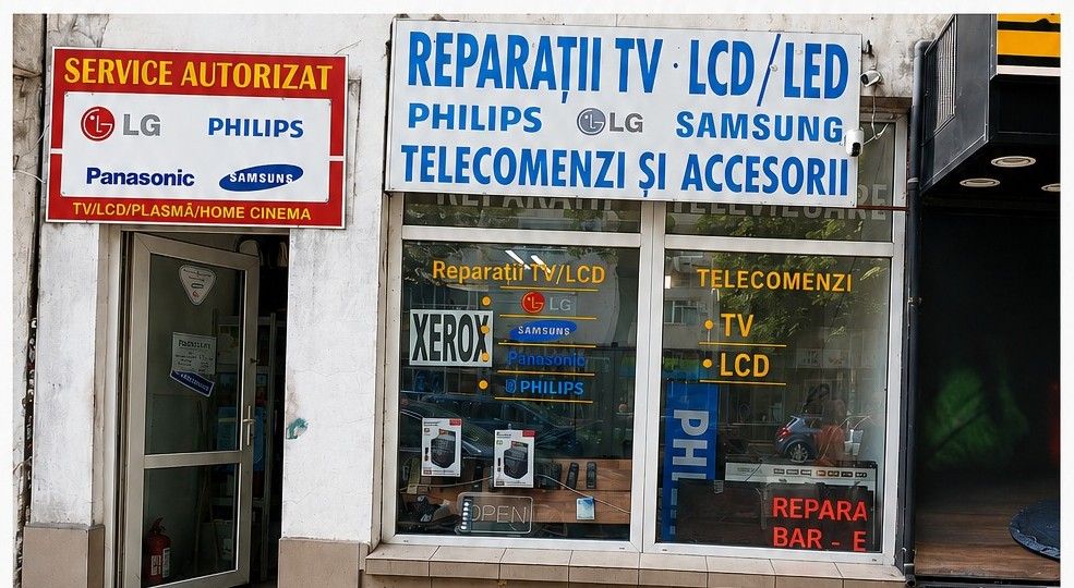 Intrarea în service: ușa albă sub panoul roșu „Service autorizat” și vitrina cu firma Reparații TV LCD / LED, telecomenzi și accesorii